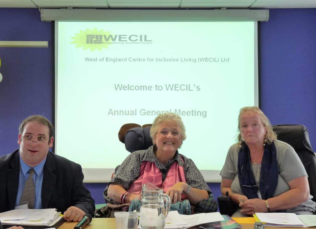 Jayne at a historical WECIL meeting, 3 people are sitting at a table with a projection screen behind them