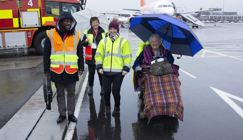 Jayne on an access audit, four people are on Bristol Airport runway, three of them have hi vis jackets, Jayne has an blue umbrella