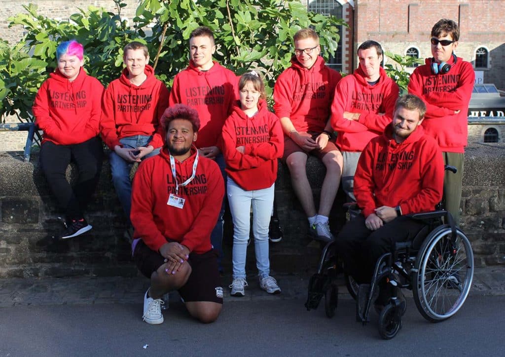 a group of young people in red jumpers which say 'listening partnership', some with visible disabilities, are standing by a wall in castle park in Bristol. The sun is shining and everyone looks happy