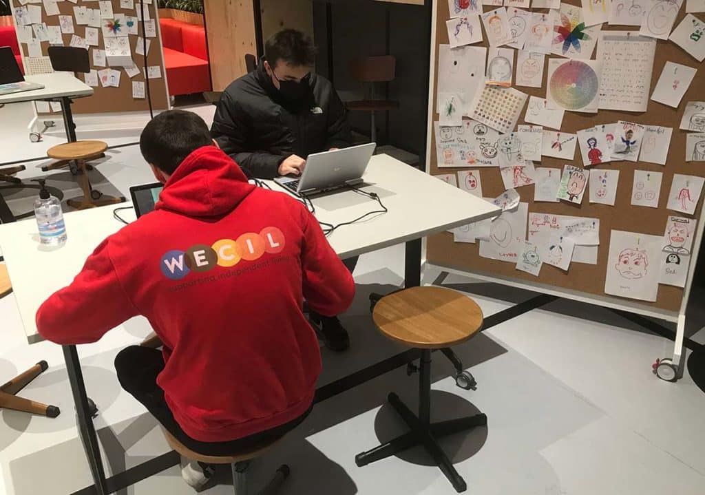 Two people are sat at a white desk, both are on laptops. The individual at the front of the picture is wearing a red jumper with the WECIL logo on. There is a cork wall to the right of the image with lots of hand drawn pictures of people pinned to it.